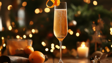 A tall champagne flute with golden-orange sparkling cocktail, garnished with a thin orange twist, set on a polished wood bar with soft warm lighting.