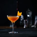 Glass of orange cocktail decorated with orange at bar counter background.