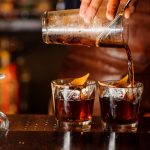 Bartender pouring fresh alcoholic drink into the glasses with ice cubes on the bar counter