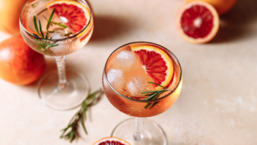 On a beige sandy background, a summer cocktail featuring blood orange, grapefruit, and rosemary. fashionable sunlight and shadows. top view, copy space, and close-up in a flat lay.