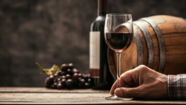 a glass of merlot in front of a barrel, bottle, and bunch of grapes