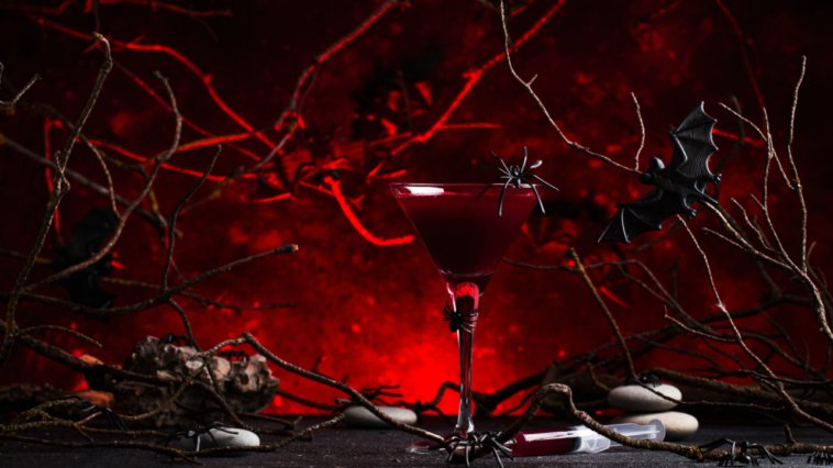 Halloween alcoholic cocktail bloody martini on scary dark red background with twisted branches, bats, stones, pumpkin guards and spiders, festive drink for vampire party