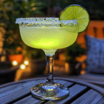Classic Margarita in a salted-rim glass with lime wheel, glowing green on a patio table with plants, cozy seating, and warm evening lighting in the background.
