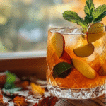 A spiced rum cocktail with apple slices and mint sits by a window, surrounded by pumpkins, apples, and fall leaves with a scenic autumn view.