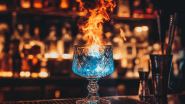 Vibrant flaming cocktail in a crystal glass with blue and orange flames rising above, set on a sleek bar surrounded by warm lights and shelves of bottles.