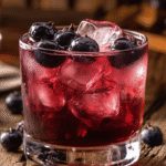 A vibrant huckleberry cocktail served over ice in a short glass, garnished with fresh huckleberries, on a rustic wooden table with a bar background.