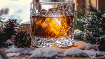 Golden brown bourbon cocktail with ice in a crystal glass, surrounded by pinecones, evergreen branches, and snow by a frosty window in a cozy winter scene.