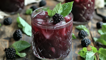 Dark purple blackberry mocktail in a textured glass with ice, garnished with a fresh blackberry and mint, surrounded by scattered berries and leaves.