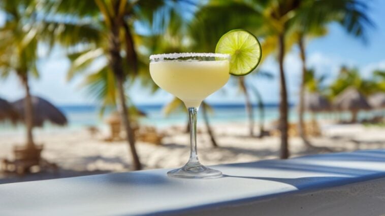 16 Must-Try Frozen Tequila Drinks 1 Frozen lime margarita in a stemmed glass with salted rim, lime wheel, and umbrella on a beach bar, with palm trees, sand, and blue sky in the background.