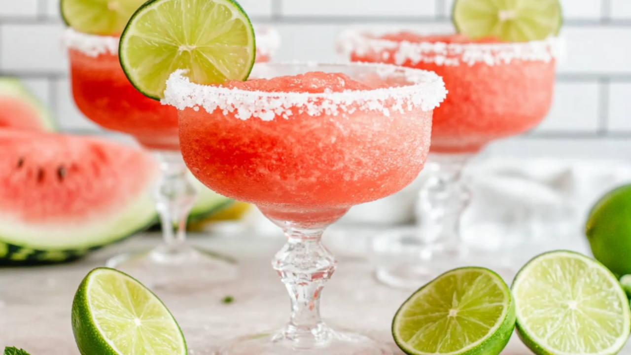 Bright pink frozen watermelon cocktails with salted rims sit in elegant glasses, surrounded by fresh watermelon slices, lime wedges, and mint leaves.