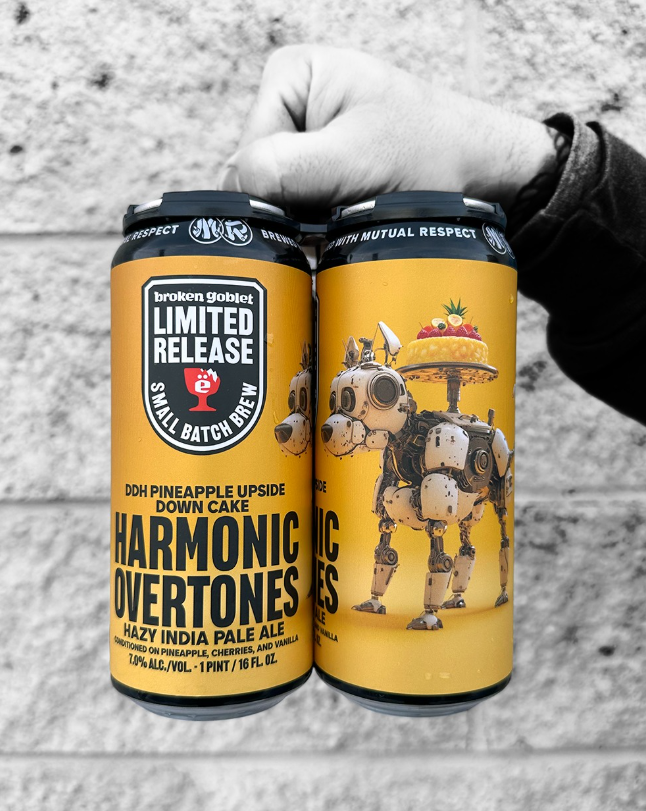 A hand holds two bright yellow cans of Broken Goblet Brewing's Harmonic Overtones hazy IPA, featuring a robot dog with a pineapple upside-down cake on its back.








