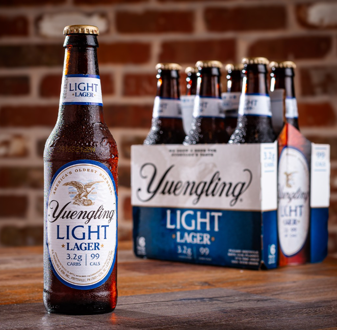 A chilled bottle of Yuengling Light Lager stands in front of a six-pack on a wooden table, with a brick wall background highlighting its crisp, low-calorie appeal.