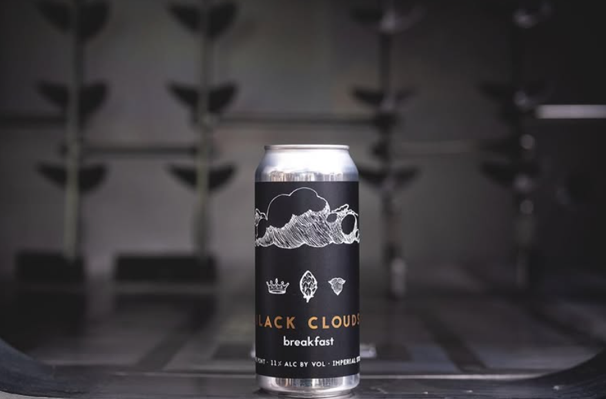 A can of Dancing Gnome's Black Clouds Breakfast Imperial Stout with a dark label featuring hand-drawn clouds, a hop, and crown icons on a metal surface.