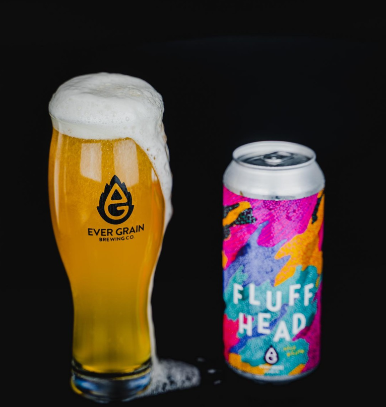A tall foamy glass of Ever Grain Brewing Co.'s Fluff Head sits next to a colorful can with bold, abstract artwork against a dramatic black background.