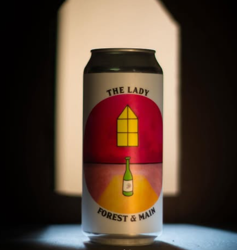 A can of The Lady by Forest & Main features surreal artwork of a green bottle lit by window light on a red and yellow background, spotlighted in the dark.