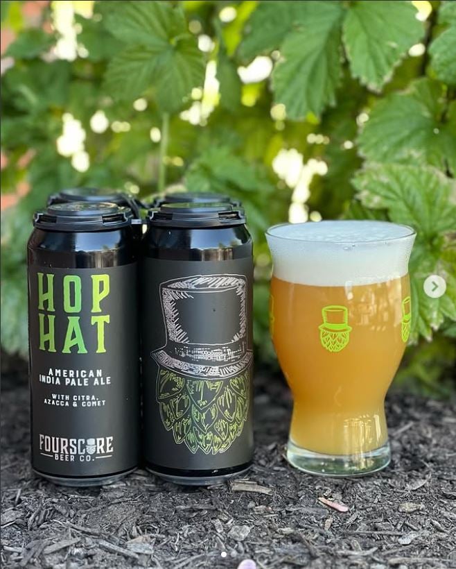 Four black cans of Fourscore Beer Co.'s Hop Hat IPA and a glass of hazy beer with frothy head are placed outdoors against green hop leaves.





