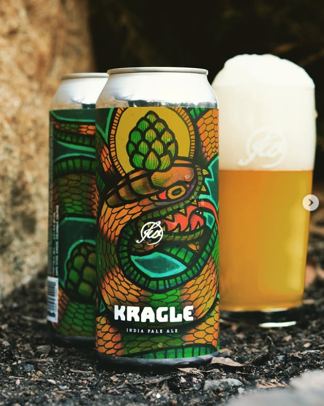 Two cans of Kragle IPA from Free Will Brewing Co. with bold serpent and hop artwork sit beside a foamy glass of hazy beer on earthy ground near a rock.