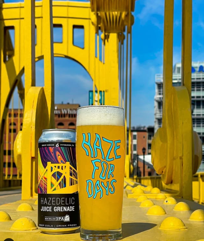 A can and glass of Grist House's Hazedelic Juice Grenade IPA sit on a bright yellow Pittsburgh bridge, with "Haze for Days" text and city buildings in the background.