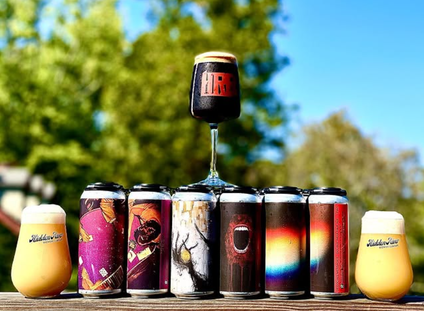 A lineup of Hidden River Brewing Company beer cans with vibrant, artistic labels, flanked by two full glasses and a stout in a goblet balanced on top.