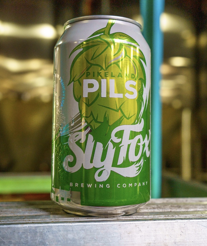 A can of Sly Fox Brewing Company’s Pikeland Pils stands on a metal surface, featuring a bold green hop design with a brewery backdrop softly blurred.