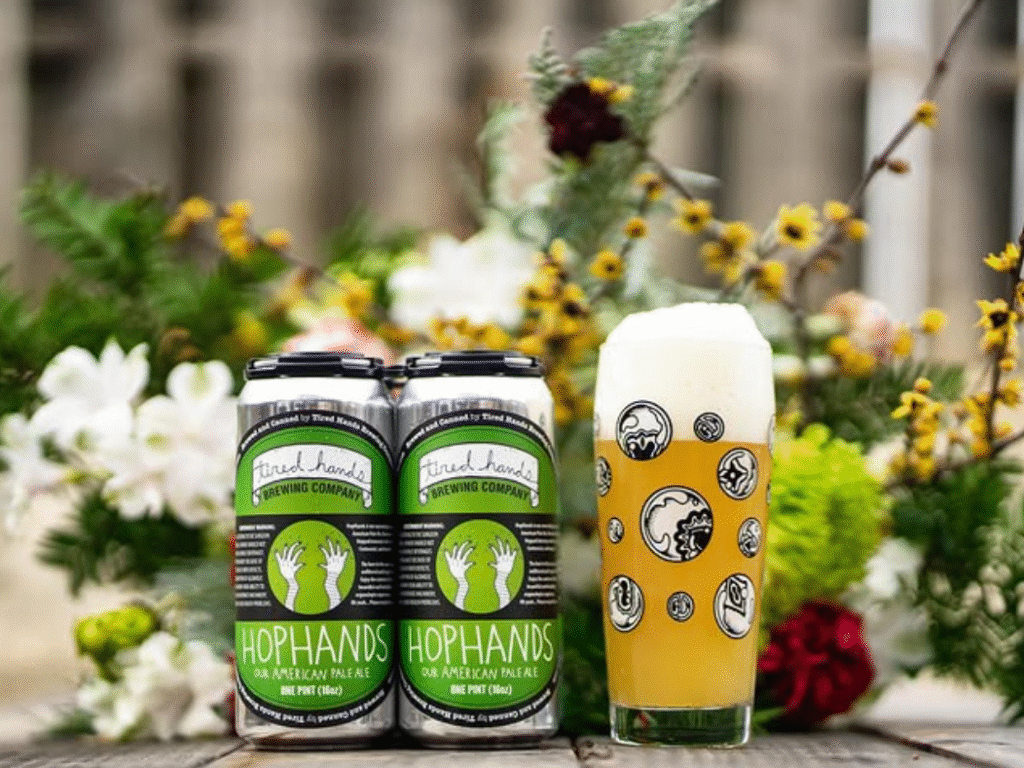 Two cans of Tired Hands Brewing Company's HopHands American Pale Ale beside a full pint glass with foamy head, set against a floral background outdoors.