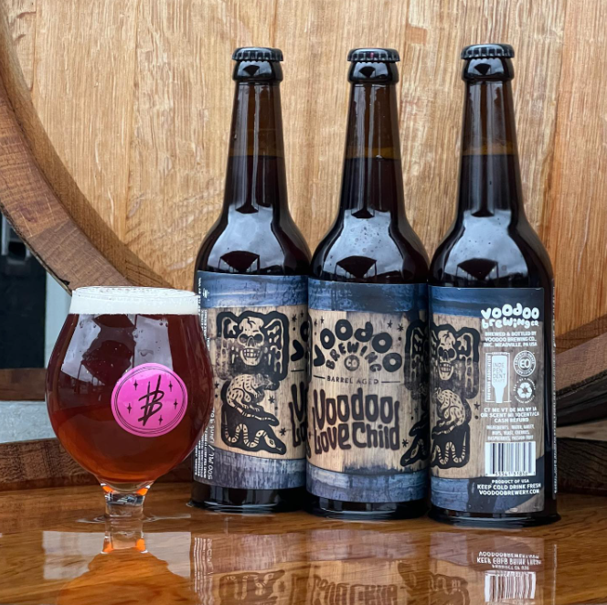Three bottles of Voodoo Brewing Co.'s Voodoo Love Child Belgian-style ale sit beside a tulip glass filled with amber beer, set against a large wooden barrel.