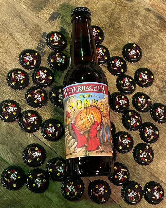 A bottle of Weyerbacher Merry Monks Belgian-style tripel lies on a wooden surface surrounded by bottle caps featuring a cheerful cartoon monk holding a beer.